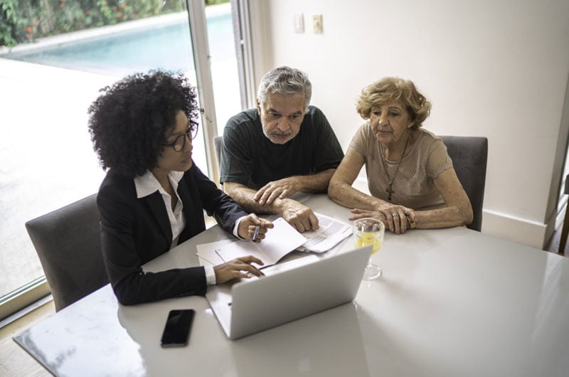 Photo of elderly couple looking at computer with younger woman with paperwork for estate planning.