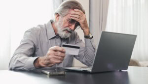 Older man in front of laptop holding a credit card and holding his head.