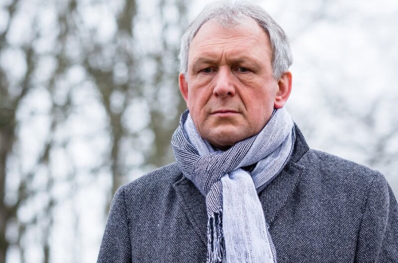 Older man walking outside, trees are leafless and he looks sad