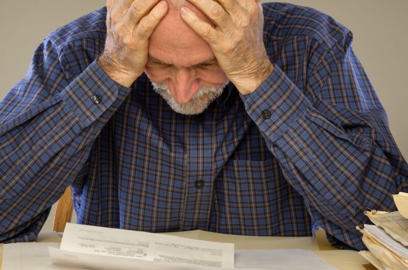 older man with hands clutching head looking at stack of bills